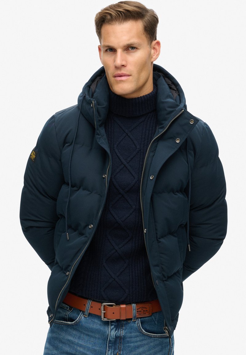 Giubbotto imbottito blu navy con cappuccio, design trapuntato, chiusura frontale con zip e bottoni a pressione. Indossato sopra un maglione blu navy a trecce e jeans blu. Cintura marrone.