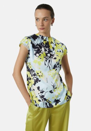 Blümchenbluse in Hellblau, mit schwarzen und gelben Mustern, kurzen Puffärmeln und einem gerafften Ausschnitt. Kombiniert mit grünen Hosen.