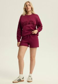 Maroon sweatshirt and shorts set with a black graphic print. Soft, textured fabric with a relaxed fit. White socks and casual shoes complete the look.