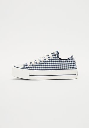 Zapatilla de cuadros de gingham en azul marino y blanco con diseño de cordones, puntera de goma blanca, suela gruesa y ribete en contraste alrededor de la base.