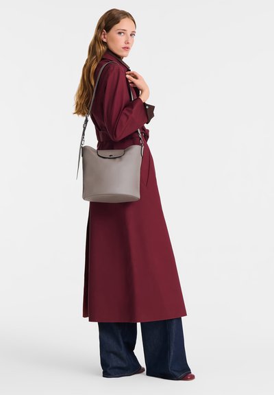 Frau in einem langen, burgunderfarbenen Mantel und weit geschnittenen Jeans, die eine graue Leder-Umhängetasche trägt, vor einem schlichten weißen Hintergrund stehend.