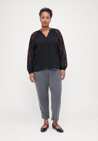 Svart bluse med spissarmer, knappfront, kombinert med grå, strukturert tapered bukser og svarte flate sko. Enfarget bakgrunn.