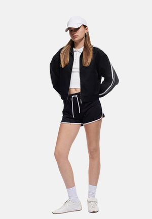 Chaqueta negra corta con rayas blancas, camiseta polo blanca, pantalones cortos negros con ribete blanco, gorra blanca y zapatillas blancas con suelas texturizadas.