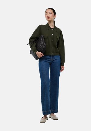 Veste tricotée vert forêt avec de grands boutons, deux poches avant, associée à un jean évasé blue et des chaussures beiges, portant un sac tressé noir.