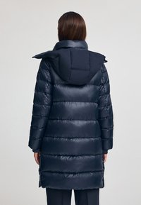 Granatowy ocieplany płaszcz z odpinanym kapturem, z pikowanym wzorem i ściągaczami na rękawach, widziany z tyłu. Miękka, błyszcząca powierzchnia.