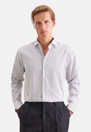 Homme portant une chemise blanche et grise à rayures verticales boutonnée et un pantalon gris foncé, les mains dans les poches, regardant devant avec une expression neutre.