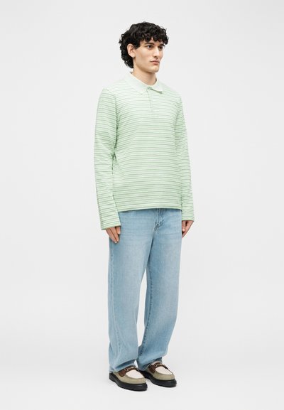 Νέος άνδρας που στέκεται φορώντας πουκάμισο πόλο μακρυμάνικο με πράσινες και λευκές ρίγες, ανοιχτόχρωμα μπλε τζιν και καφέ με μπεζ μοκασίνια.