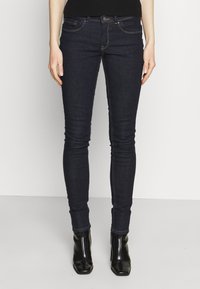 Jeans skinny in denim scuro con una texture liscia. Presentano cuciture a contrasto, tasche anteriori e una silhouette aderente, abbinati a tacchi neri.