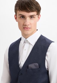 Granatowa kamizelka o fakturze z klapami typu notch, noszona na białej koszuli z guzikami. Zawiera poszetkę w wzorzystym materiale.