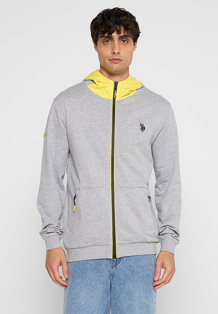 Sweat-shirt zippé gris clair avec une capuche intérieure jaune, une fermeture à glissière noire, des poches latérales et un logo brodé sur la poitrine. Texture de tissu douce.