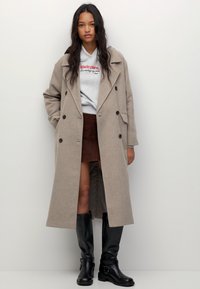 Femme portant un manteau long beige par-dessus un hoodie gris, une jupe marron et des bottes noires hautes, se tenant devant un fond clair uni.