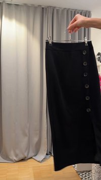 Hånd, der holder et sort nederdel med seks knapper ned langs siden, vist på en gennemsigtig bøjle i et prøverum.