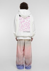 Felpa bianca con una grafica rosa ondulata e testo sul retro, abbinata a pantaloni larghi oversize in gradiente pastello che vanno dal rosa al blu.