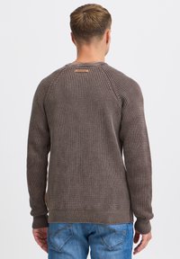 Brązowy sweter o teksturowej strukturze z rękawami raglanowymi, żebrowanymi mankietami i dołem, z małą metką marki z tyłu. W połączeniu z niebieskimi dżinsami.