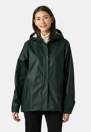 Mujer con chaqueta de lluvia con capucha de color verde oscuro y un jersey de cuello alto claro, de pie frente a un fondo claro y liso, sonriendo.