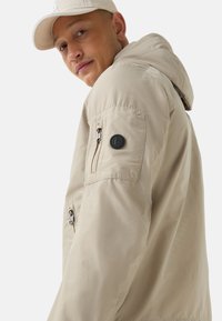 Beige hooded jacket with a smooth finish, featuring a side pocket, zipper closure, and a branded black circular accent on the sleeve.