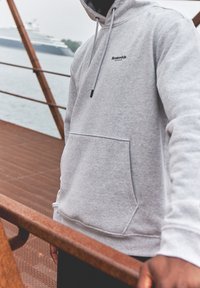 Hoodie harmaa puuvillasta, jossa on säädettävä nyöri, etutasku ja logoyksityiskohta. Tekstuuri vaikuttaa pehmeältä; ympäristönä on vesiraja kaiteineen.