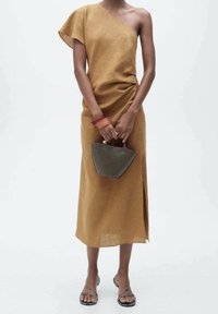 Asymmetrisches Ein-Schulter-Kleid aus senfgelbem Stoff mit einem seitlichen Schlitz. Schlichtes Design kombiniert mit einer kleinen grauen Handtasche und Riemchensandalen.