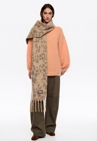 Beżowy szalik w panterkę z frędzlami, wykonany z miękkiego materiału, zarzucony na różowy, miękki sweter oraz oliwkowo zielone spodnie.