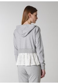 Grå hoodie med en vit broderad spetspeplum längst ner, designad med en mjuk textur, justerbar huva och ribbad midja.