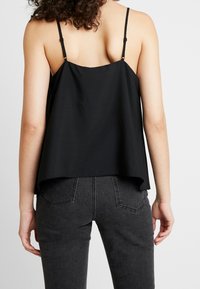 Top negro sin mangas con un corte holgado, hecho de una tela suave. Presenta tirantes finos ajustables y un dobladillo curvado, combinado con jeans gris oscuro.