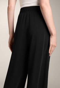 Czarne spodnie o szerokich nogawkach z gładką teksturą, elastycznym pasem i luźnym krojem, pokazane od tyłu z podkreśleniem bocznego szwu.