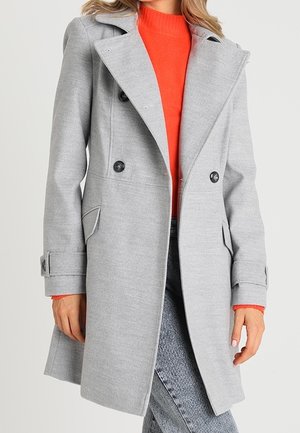 Femme portant un manteau gris clair à double boutonnage sur un pull orange avec un jean bleu clair.