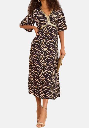 Mujer con un vestido midi negro con rayas onduladas amarillas y rosas, ribete dorado en el escote, sosteniendo un bolso de mano marrón y llevando tacones beige.