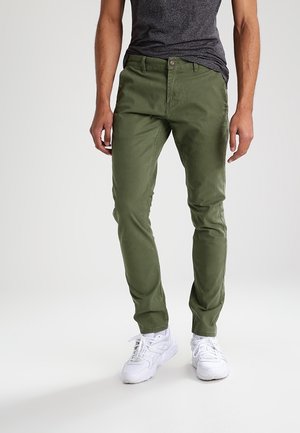 Mężczyzna w oliwkowozielonych spodniach, ciemnoszarej koszulce i białych trampkach stojący na tle jasnego, jednolitego tła.