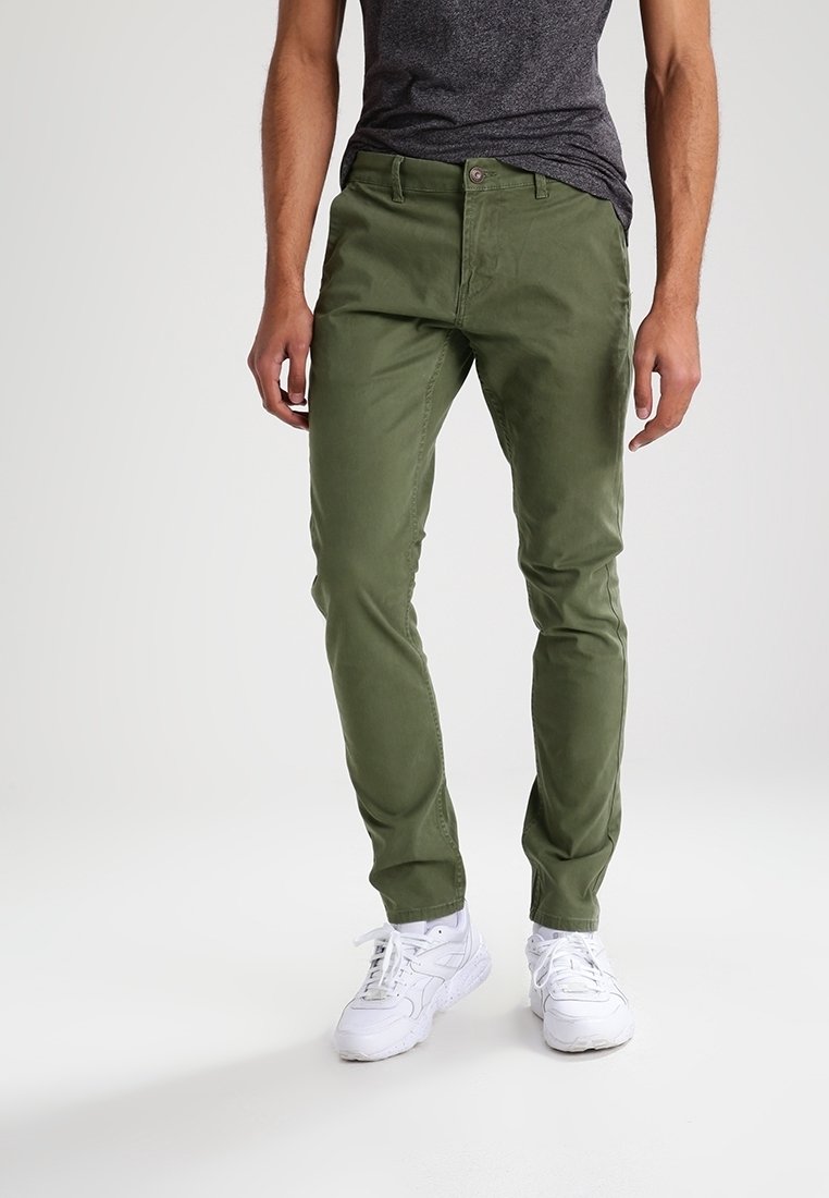 Homme portant un pantalon vert olive, un t-shirt gris foncé et des baskets blanches, debout devant un fond clair uni.
