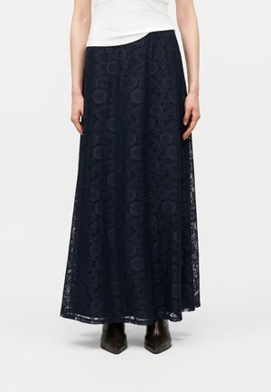 Žena v tmavomodrej maxi sukni s kvetinovou čipkou, bielom top-e a čiernych špicatých čižmách stojí na jednoduchom svetlom pozadí.