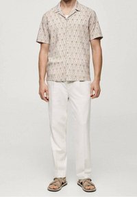 Koszula z krótkim rękawem w beżowym kolorze z geometrycznymi wzorami, zestawiona z białymi lnianymi spodniami i szarymi sandałami.