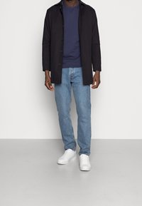 Abrigo negro de botones sobre un sudadera azul marino, combinado con jeans azul claro y zapatillas blancas. Diseño sencillo sin patrones o acentos visibles.