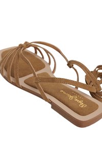 Brun sandal i mocka med platt sula, öppen tå och flera korsande remmar. Har en vadderad fotbädd med logotypdetalj.