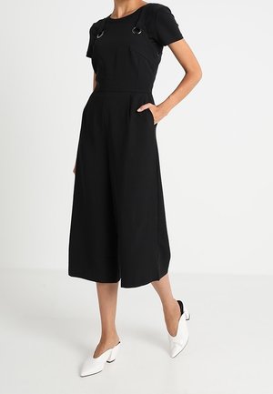 Kvinde iført sort bredbenet cropped jumpsuit med skulderringdetaljer og hvide mule-sko med spidst tåpåkryds, stående med den ene hånd i lommen.
