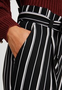 Černobílé pruhované kalhoty s vysokým pasem, disponující bočními kapsami a strukturovaným materiálem. Ozdobené detailem s páskem v pase.