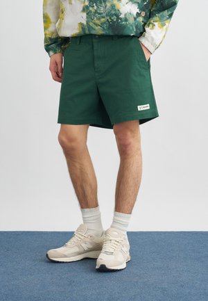 Person som har på seg mørkegrønne shorts, hvite sokker, beige joggesko og en langermet skjorte med grønt-gult mønster, står på blått teppe.