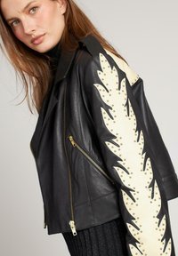 Vrouw die een zwart leren jack draagt met een gouden rits en crèmekleurige studs in de vorm van vlammen langs de mouwen, poserend tegen een neutrale achtergrond.