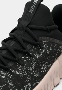 Κοντινό πλάνο μαύρου αθλητικού παπουτσιού με λευκό σπαστό σχέδιο, μαύρες κορδόνες, μαλακή γλώσσα και ανοιχτό μπεζ υφαντή σόλα.