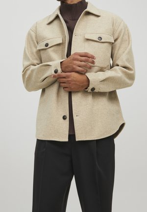 Personne portant une veste-chemise en laine beige boutonnée sur un col roulé marron à côtes, ajustant le poignet, associée à un pantalon noir ample.