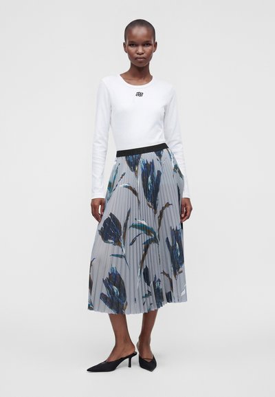 Mannequin portant un haut blanc à manches longues et une jupe midi plissée à motifs floraux bleus, associée à des chaussures noires à talons avec bride arrière.