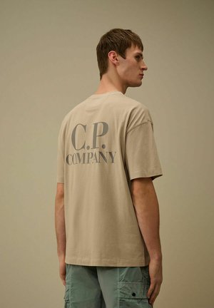 Giovane uomo con t-shirt beige C.P. Company e pantaloncini cargo verdi, in piedi di lato su uno sfondo beige uniforme.