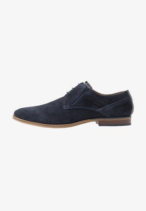 Chaussure de ville lacée en daim bleu marine pour homme, avec semelle marron clair, talon bas empilé, détails de coutures latérales et marquage embossé.