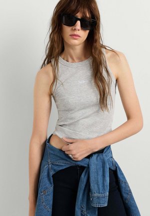 Junge Frau mit schwarzen Sonnenbrillen, einem hellgrauen ärmellosen Top und einer Jeansjacke, die um die Taille gebunden ist, vor einem einfarbigen Hintergrund.
