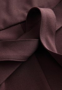 Tissu en daim brun foncé avec des coutures visibles, plié et bouclé, mettant en valeur sa texture douce et son savoir-faire détaillé.