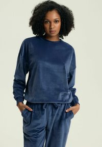 Granatowy welurowy sweter z długimi rękawami, okrągłym dekoltem i luźnym krojem. Posiada kieszenie i gładką fakturę. Zestawiony z pasującymi spodniami.