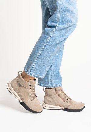 Persona con botines de ante beige con suelas negras y vaqueros de mezclilla azul claro sobre un fondo blanco.