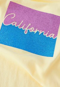 Bawełniana koszulka z żółtym tłem, na którym znajduje się prostokąt z gradientem brokatowym w kolorach purpurowym i niebieskim, z napisem "California" w białej, kursywowej czcionce.