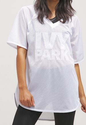 Osoba nosząca białą perforowaną, oversize'ową koszulę z napisem "IVY PARK" dużymi literami z przodu, w połączeniu z czarnymi legginsami.