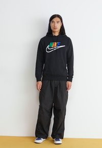 Felpa nera con logo Nike colorato, abbinata a pantaloni cargo neri larghi e sneakers bianche con accenti blu. Texture liscia.